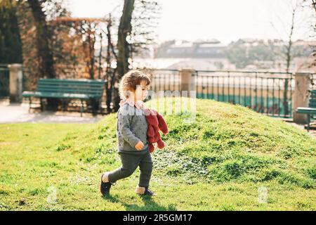 Outdoor portrait of adorable toddler girl playing with pink bunny toy in sunny green park in early spring Stock Photo