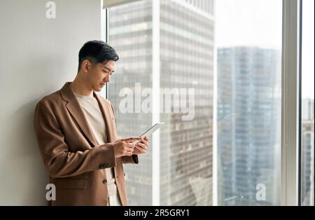 Professional young Asian businessman using tablet standing at window. Stock Photo