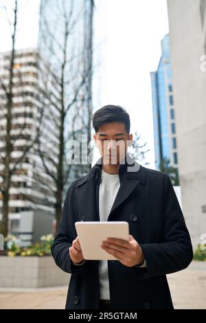 Young Asian business man standing in city using digital tablet. Vertical Stock Photo