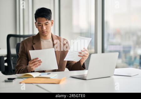 Young busy Asian business man holding documents sitting at desk in office. Stock Photo