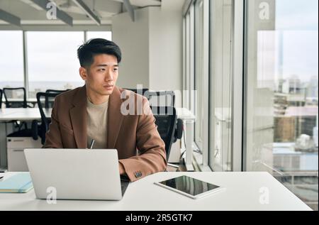 Young Asian businessman looking away thinking working on laptop in office. Stock Photo