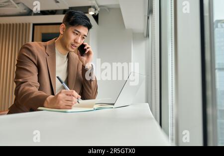 Busy Asian business man making phone call using laptop in office. Stock Photo