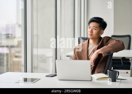 Young Asian business man using laptop looking away thinking in office. Stock Photo