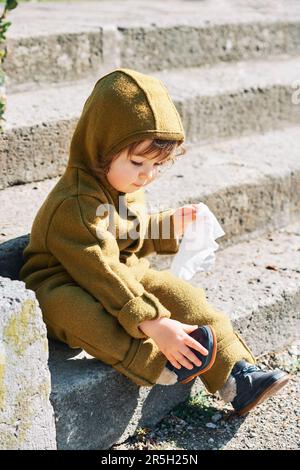 Cute toddler kid cleaning shoes with wet wipe, sitting on steps in public park, wearing green warm jumpsuit Stock Photo