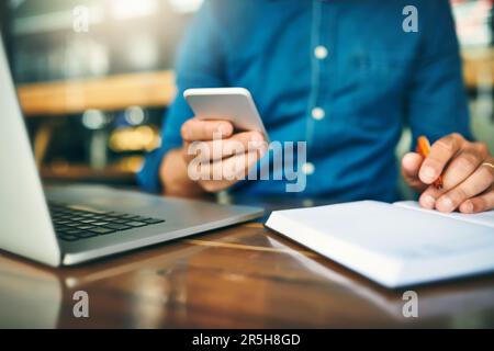 Staying connected has never been easier. an unrecognizable businessman sitting alone and making notes while using his cellphone in a cafe. Stock Photo