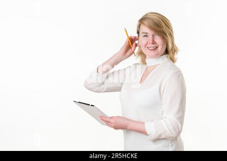 Smiling female teacher straightens blond hair on head with hand with pencil. Happiness woman holding clipboard and looking at camera. Blonde woman Stock Photo