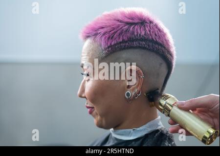 The hairdresser shaves the temple of a female client. Asian woman with short pink hair in barbershop. Stock Photo