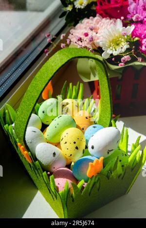 Easter green basket with plastic colored eggs on the windowsill against the background of flowers Stock Photo