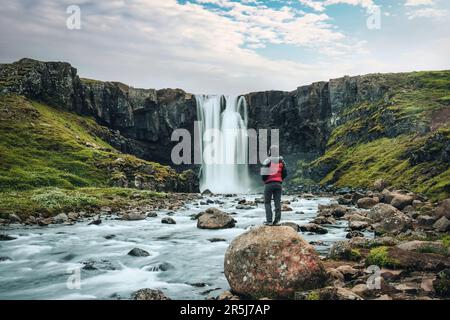 Beautiful view of Gufufoss waterfall flowing with young asian tourist woman standing on the rock in summer at Seydisfjordur, East of Iceland Stock Photo
