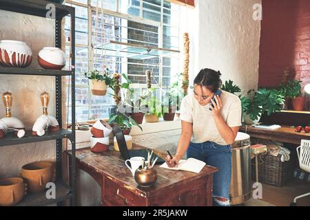 Im making a note of your order. an attractive young business owner standing and using her cellphone while taking notes in her pottery studio. Stock Photo