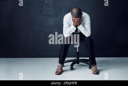 Everyone has battles to face before they see success. Studio shot of a young businessman sitting with his hands covering his face against a dark Stock Photo