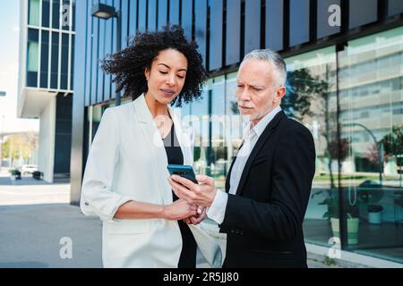 Handsome businessman wearing suit showing information to his colleague woman on a smartphone standing at the office building close to work company. Pair of executive people watching a mobile phone. High quality photo Stock Photo