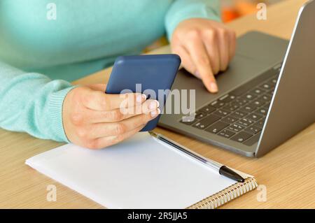 Close up of a woman hands using a smart phone and a laptop on a desktop at home or office Stock Photo