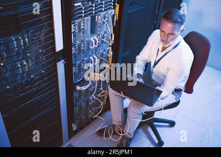 Hes the go-to person for technical problems. High angle shot of a mature man using a laptop while working in a server room. Stock Photo