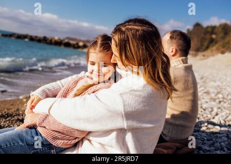 Man, woman and little child by the sea. Mom, dad and female  sitting back. Cute little girl smiling and looking at camera. People are dressed in warm Stock Photo