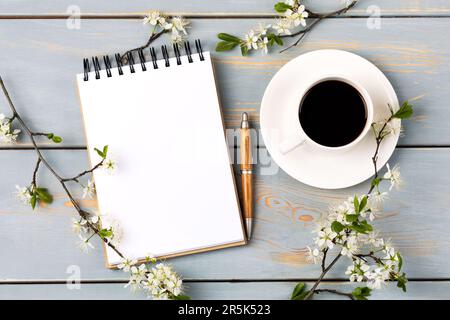 Artistic composition with opened blank notepad and pen, cup of black coffee, branch of white cherry blossoms on blue wooden table. Festive office desk Stock Photo