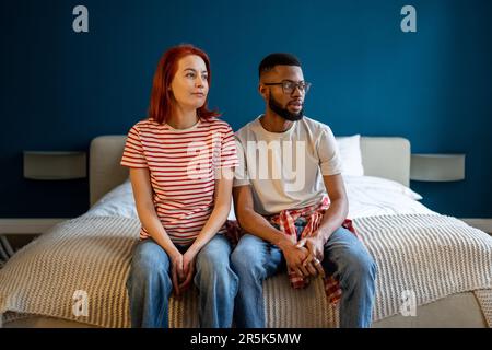 Pensive multiracial family couple sitting on comfortable bed daydreaming together looking to window. Stock Photo