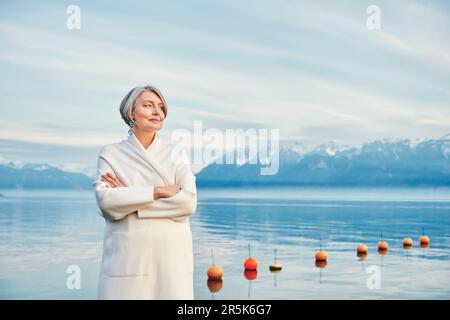 Outdoor portrait of beautiful middle age woman posing next to winter lake, wearing white coat Stock Photo