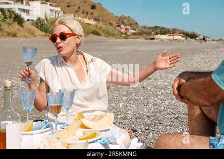 Summer beach picnic. Middle aged couple having picnic with fresh fruit, wine and appetizers outdoors at seaside at the beach. Blond woman having fun w Stock Photo