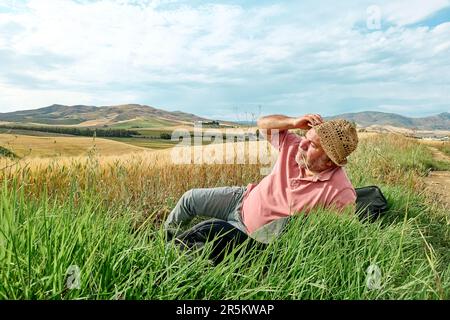 Middle-aged man resting in poppies meadow near golden wheat field in hot summer sun and blue sky with white clouds with mountains hill landscape in ba Stock Photo