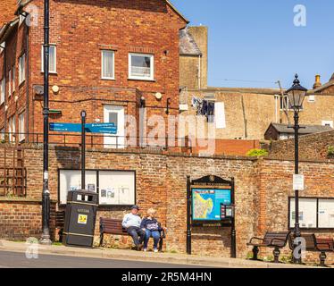 Two people sit on a sloping bench beside a steep road. A lamppost and signpost are on either side and a house is behind with washing on a line. A town Stock Photo