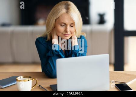 Exhausted mature businesswoman working on laptop and rubbing neck, suffering from office syndrome, home interior Stock Photo