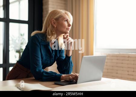 Pensive mature woman entrepreneur pondering business ideas while working at home, leaning chin on hand and looking away Stock Photo