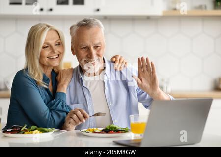 Happy senior spouses making video call with laptop in kitchen during breakfast Stock Photo