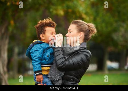 Young mother holding crying toddler, parent in park with sad child, mix race family Stock Photo