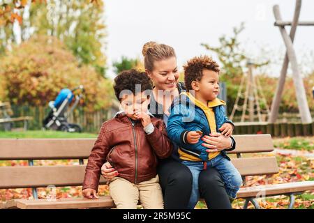 Outdoor portrait of happy young mother with two lovely sons, family enjoying nice autumn day in public park, mixed race family. Kids eating snacks, re Stock Photo