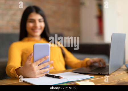 Educational gadgets. Happy student lady using cellphone, sitting with laptop at desk, learning online holding smartphone Stock Photo