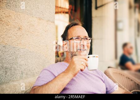 Happy laughing man relaxing in summer outdoor cafe, holding cup of coffee, wearing purple t-shirt and eyeglasses Stock Photo