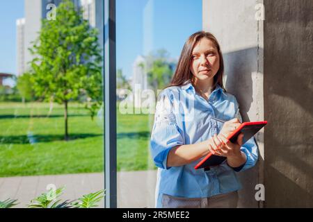 Serious confident office worker holding tablet standing by window. Caucasian young woman using modern digital technologies. Business concept. Space Stock Photo
