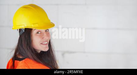 Young woman in yellow hard hat and orange high visibility vest, long dark hair, looking over her shoulder, smiling confident. Blurred construction sit Stock Photo