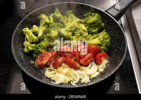 Onions, tomatoes and broccoli are sauteed in a steaming frying pan on the black stovetop for a healthy vegetarian meal, selected focus, narrow depth o Stock Photo