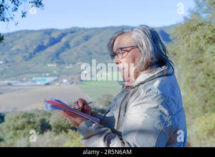 Woman with white hair and glasses writing in a notebook with a pencil in the background of a mountainous landscape Stock Photo