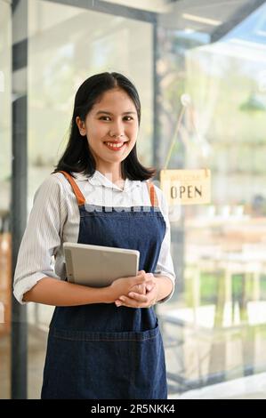 A beautiful and friendly young Asian female waitress in an apron stands at the coffee shop entrance door with her digital tablet. Hospitality service, Stock Photo