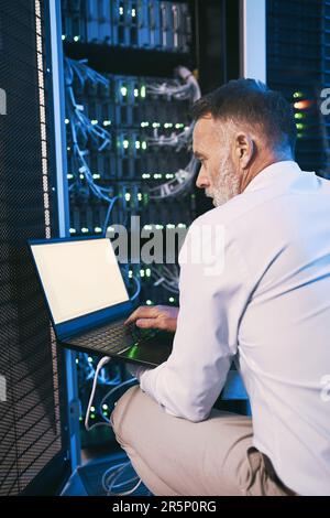 Big data is his business. Rearview shot of a mature man using a laptop while working in a server room. Stock Photo