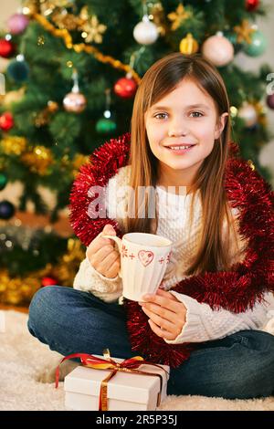 Portrait of sweet little girl sitting next to Christmas tree, wearing white cozy pullover, holding cup with hot chocolate, winter holidays Stock Photo