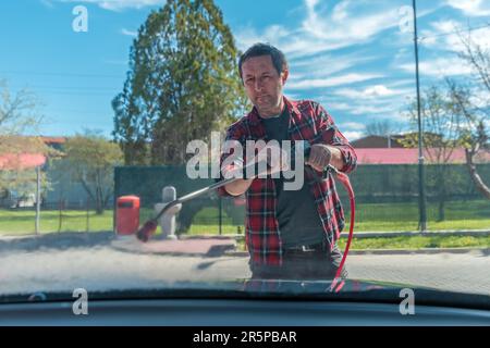 Man washing car with water gun in carwash self-service. Soap sud, wax and water drops covering vehicle window glass. Seen from the inside of the autom Stock Photo