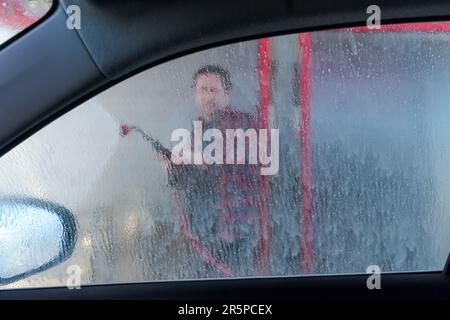 Man washing car with water gun in carwash self-service. Soap sud, wax and water drops covering vehicle window glass. Seen from the inside of the autom Stock Photo