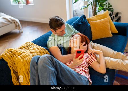 Couple man woman looking each other lying on couch with smartphone at home. Stock Photo