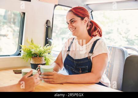 Two women drinking coffee and having a great time in a camper van. Van road trip holiday and outdoor summer adventure. Nomad lifestyle concept Stock Photo