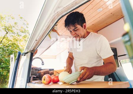 Young latin man cooking food in the kitchen of his handmade camper van. Van road trip holiday and outdoor summer adventure. Nomad lifestyle concept Stock Photo