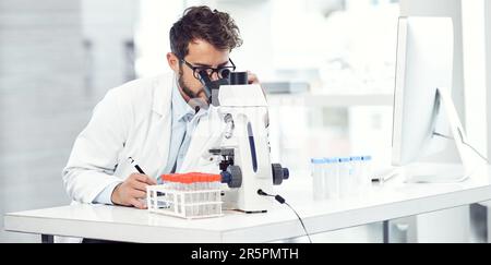 Doing what medical professionals do best. a focused young male scientist making notes while looking trough a microscope and being seated inside a Stock Photo
