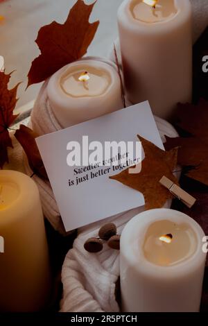 SWEATER WEATHER IS JUST BETTER TOGETHER text greeting card concept Celebrating autumn holidays at cozy home on the windowsill Hygge aesthetic atmosphere Autumn leaves spices and candle on knitted white sweater in warm yellow lights. Still life. Raining Outside Stock Photo