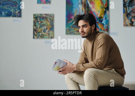 Young pensive man sitting on couch with brochure during his visit to exhibition of art gallery Stock Photo