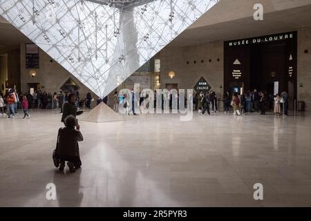 People in Paris, France gather around the captivating inverted pyramid located within the renowned shopping mall, 'Carrousel du Louvre', as people line up to enter the musuem in the background. Stock Photo