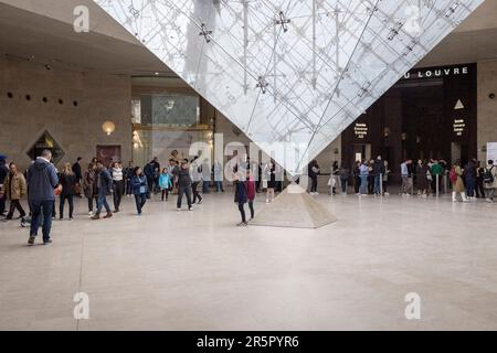 People in Paris, France gather around the captivating inverted pyramid located within the renowned shopping mall, 'Carrousel du Louvre', as people line up to enter the musuem in the background. Stock Photo