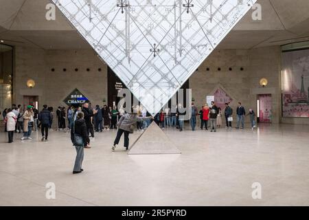 People in Paris, France gather around the captivating inverted pyramid located within the renowned shopping mall, 'Carrousel du Louvre', as people line up to enter the musuem in the background. Stock Photo